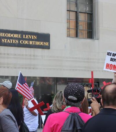 A crowd of protestors stand in front of the Theodore Levin US Courthouse holding anti-ICE protest signs and red crosses