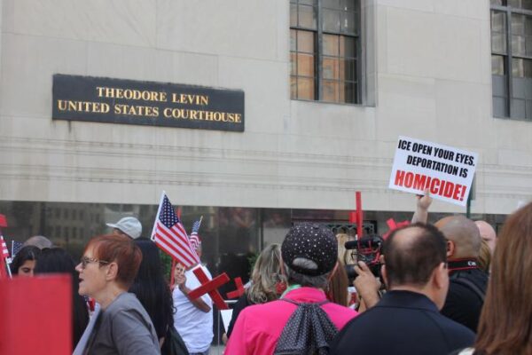 A crowd of protestors stand in front of the Theodore Levin US Courthouse holding anti-ICE protest signs and red crosses