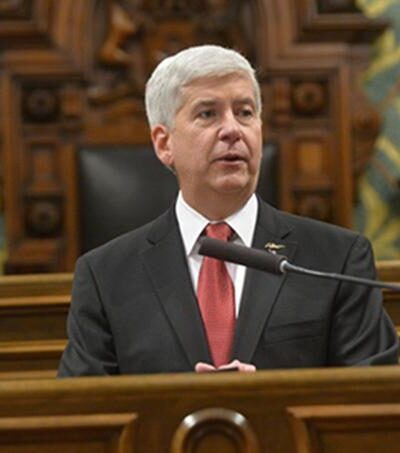 Governor Snyder sits behind a podium speaking into a microphone. He has close cropped grey hair and wears a suit with a red tie.