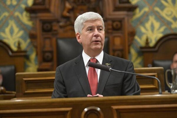Governor Snyder sits behind a podium speaking into a microphone. He has close cropped grey hair and wears a suit with a red tie.
