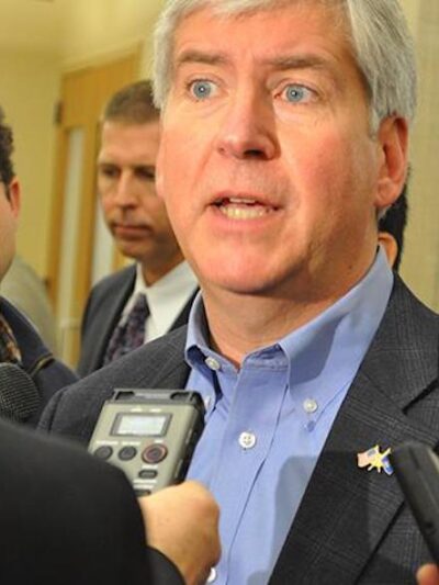 Reporters interview a man who looks stressed. He looks off into the distance as he speaks. He wears a checked suit jacket and blue collar.
