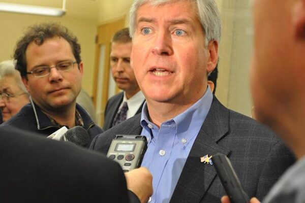 Reporters interview a man who looks stressed. He looks off into the distance as he speaks. He wears a checked suit jacket and blue collar.