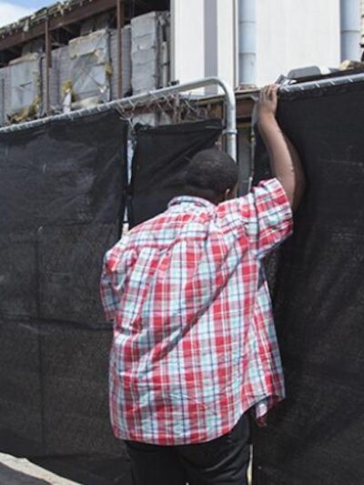 A young boy leans against a fence covered by black cloth. Behind the fence is a construction zone.