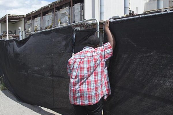 A young boy leans against a fence covered by black cloth. Behind the fence is a construction zone.