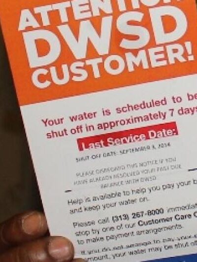 A hand holds a Detroit water department notice that says Your water is scheduled to shut off in approximately 7 days.