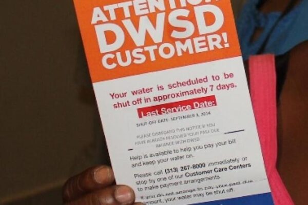 A hand holds a Detroit water department notice that says Your water is scheduled to shut off in approximately 7 days.