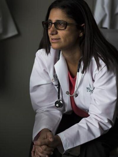 Dr. Mona Hanna-Attisha sits in a chair leaning forward and clasping her hands. She wears black rimmed glasses, a white coat, pink top and dark pants. She looks to the viewer's left.
