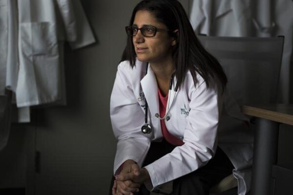 Dr. Mona Hanna-Attisha sits in a chair leaning forward and clasping her hands. She wears black rimmed glasses, a white coat, pink top and dark pants. She looks to the viewer's left.
