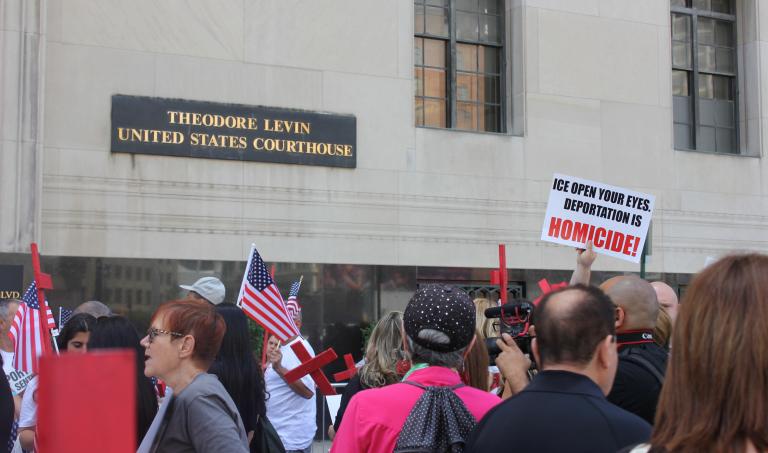 A crowd of protestors stand in front of the Theodore Levin US Courthouse holding anti-ICE protest signs and red crosses