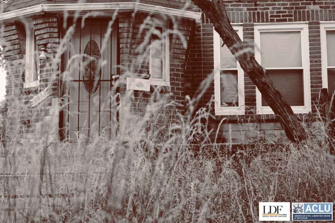 A foreclosed brick house with a black metal gate in front of the door. Weeds and grass are growing tall in the front yard. 