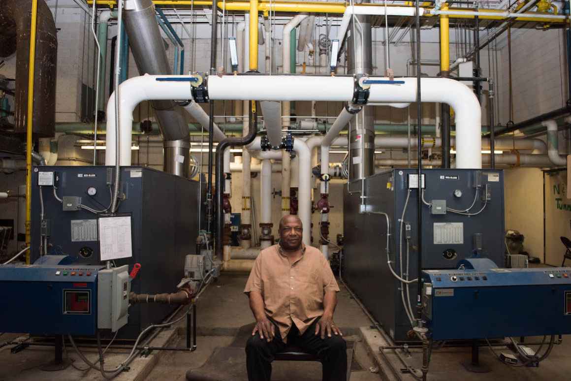 Sam Bell, a custodian at Beecher High School and General Motors retiree, sits in front of the school's antiquated steam heating system.