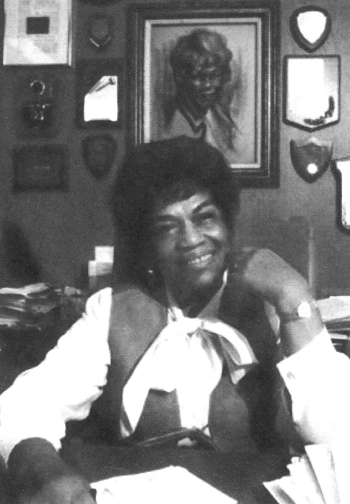 Black and white portrait of Erma Henderson sitting before a wall of portraits and plaques