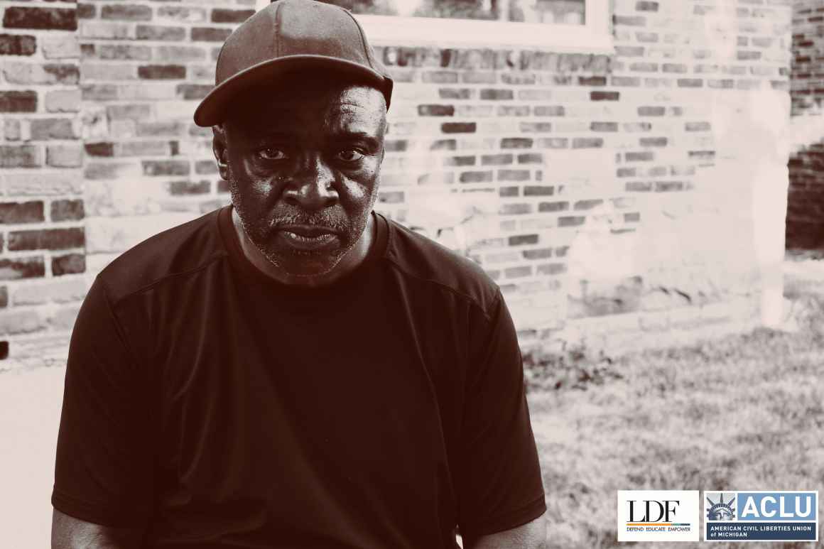 Walter Hicks sits in front of a brick wall, facing the camera. He has a serious expression and wears a baseball hat and black t-shirt. 