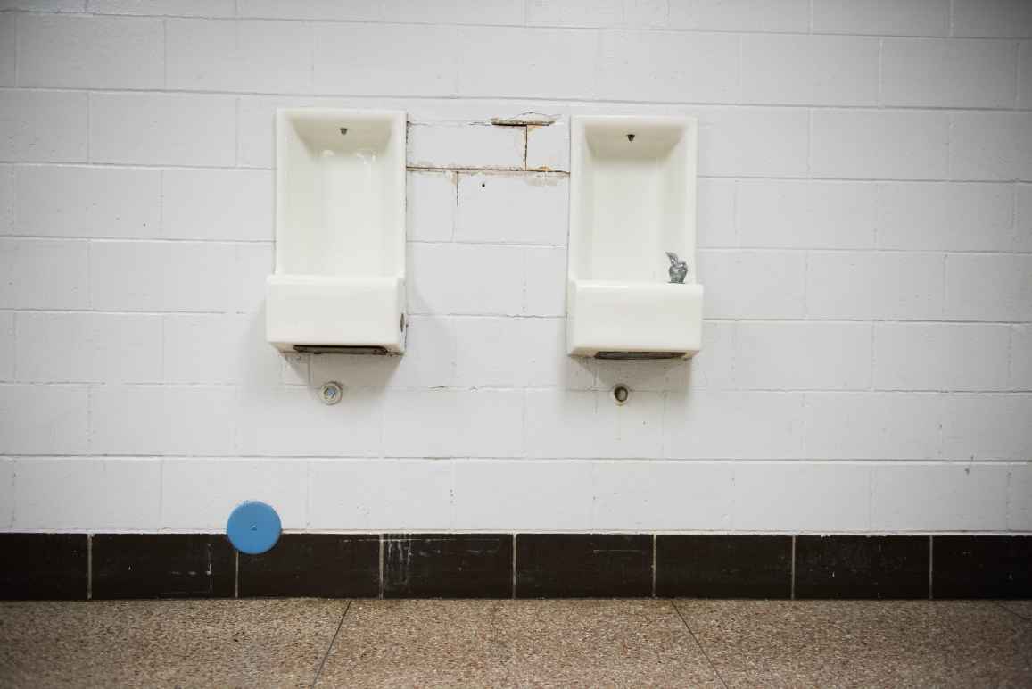 Two water fountains are in the center of a white interior brick wall. The space between the fountains is cracked and brown. Black tile runs along the bottom of the wall near the linoleum floor 