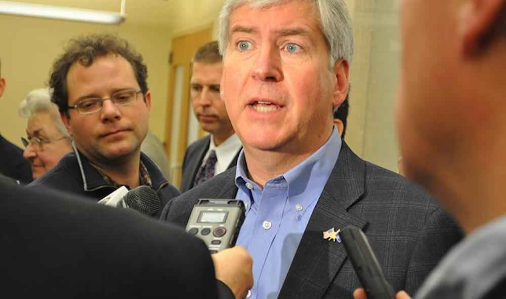 Reporters interview a man who looks stressed. He looks off into the distance as he speaks. He wears a checked suit jacket and blue collar. 