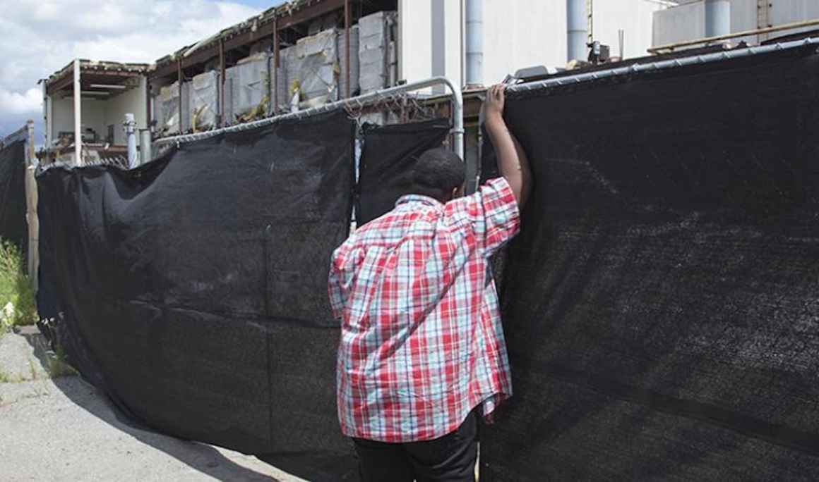 A young boy leans against a fence covered by black cloth. Behind the fence is a construction zone. 