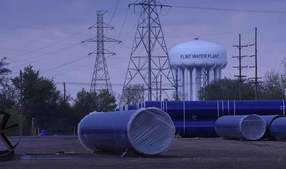 A Flint Water Plant tower against a darkened sky. In front of it are electrical wires and giant tubes on the ground. 