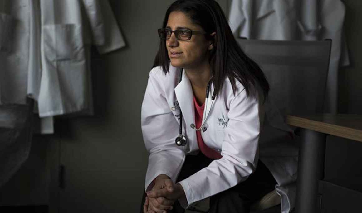 Dr. Mona Hanna-Attisha sits in a chair leaning forward and clasping her hands. She wears black rimmed glasses, a white coat, pink top and dark pants. She looks to the viewer&#039;s left. 