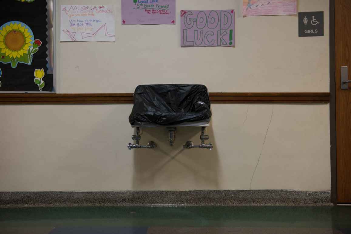 A drinking fountain covered with black plastic. Above it are hand-drawn posters wishing 5th graders good luck. On the left is a bulletin board with a black background and paper sunflower.   