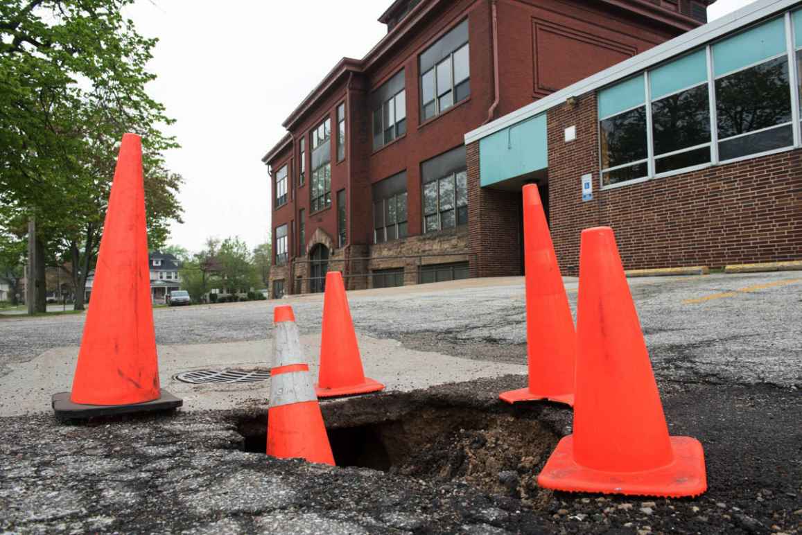 Orange traffic cones circle a hole in a road. In the background there is a red brick building on the right and a tall tree on the left