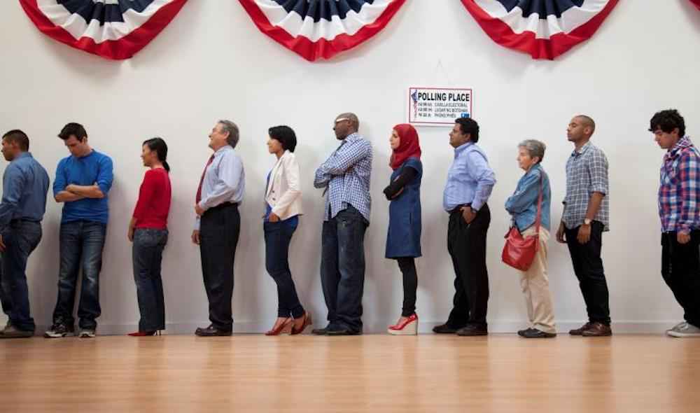 A line of people wait at their polling place beneath red, white and blue banners hanging on the wall