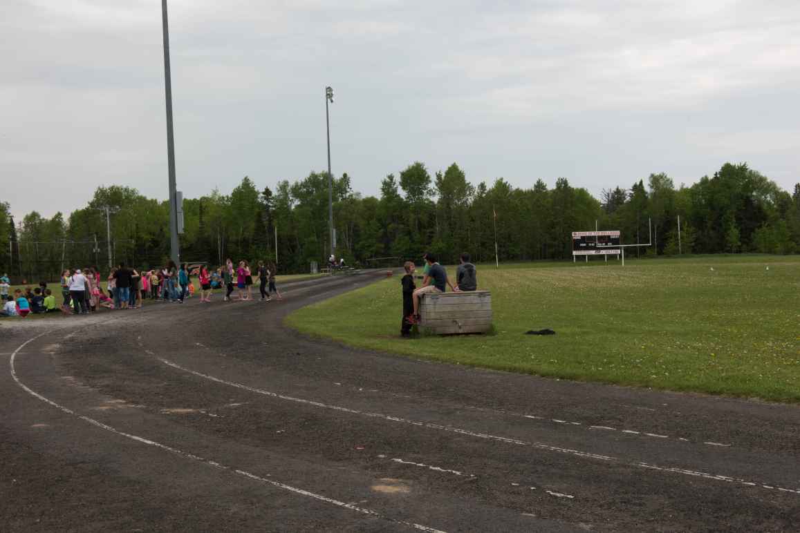 Students stand on a run-down running track. Several of the students sit on a wooden box placed on a lawn to the right.