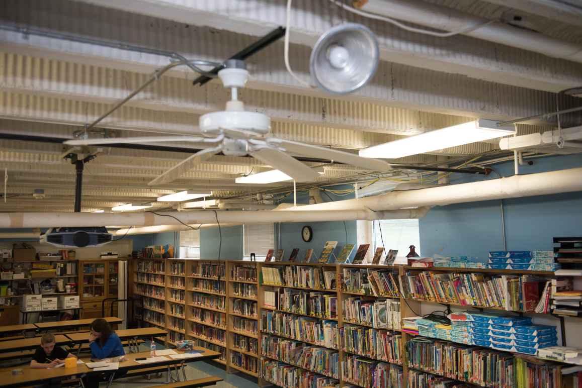 The Sodus library from above. A woman sits at a table on the left with a young boy. The shelves hold books, paper, and office equipment. The ceiling has a fan, clamp lamps, fluorescent lights and wiring. 